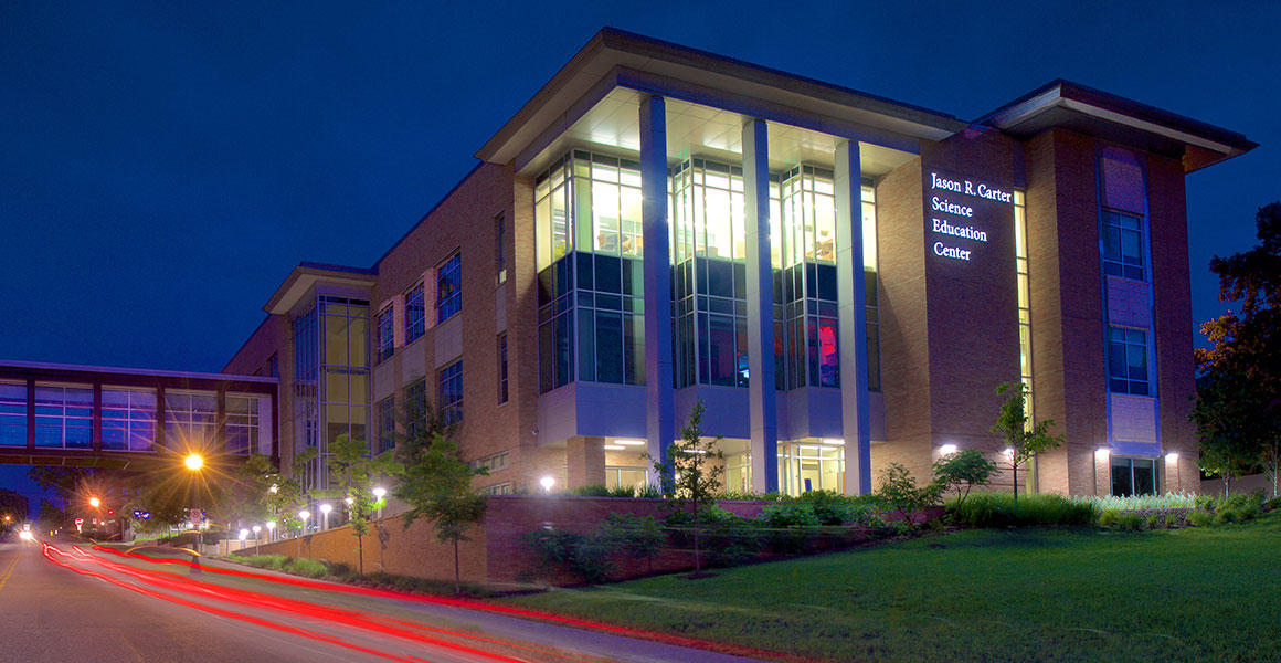 Evening photograph of Metro State University’s Jason R. Carter Science Education Center located on the Saint Paul Campus. Evening sky is dark blue with building light shining bright and remnants of taillights appearing as red streaks from cars driving by on East Sixth Street, West to East.