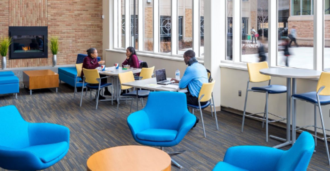 Photograph in Metro State University’s Student Center where two female students are eating their lunches and a male student studying at the next table, with a gas fireplace within a brick wall burning a low flame in an otherwise decorated room with bright blue cushioned chairs, yellow backed and dark blue table chairs, and large windows where passersby can be seen walking towards campus and into the parking ramp. 