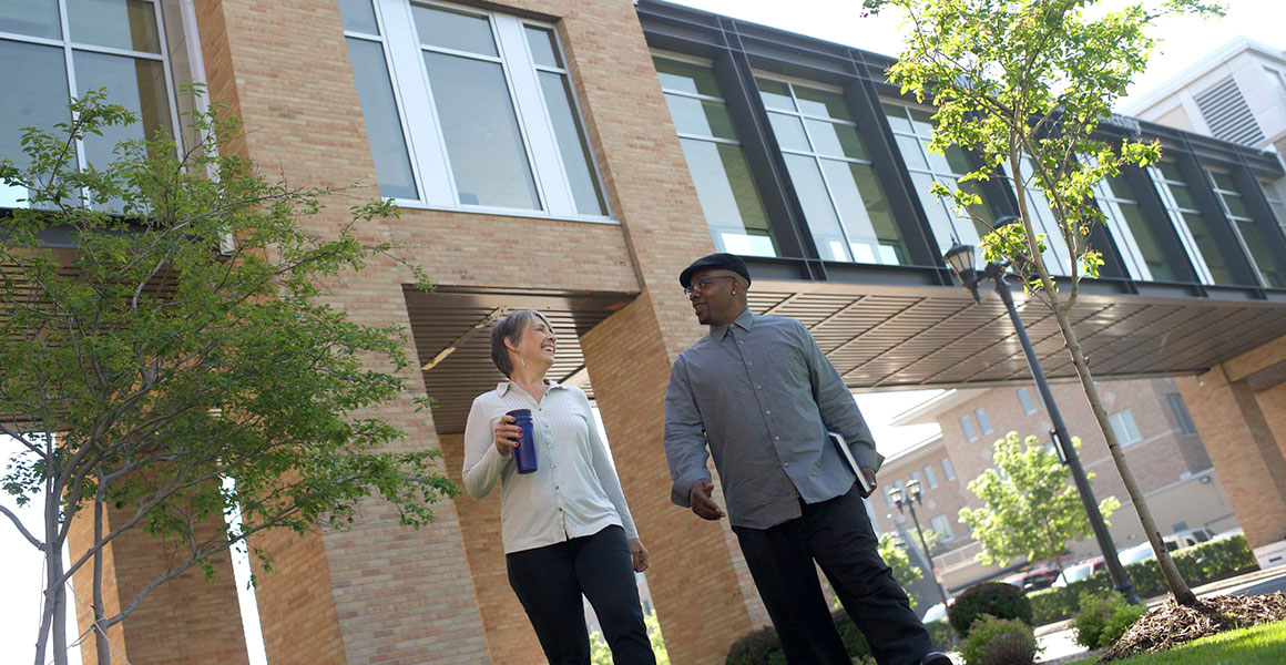 Photograph of an African American male and a Caucasian female smiling, walking and talking on the grounds of Metro State University’s library and learning center with the skyway shown that connects the library to the other building on the Saint Paul campus.