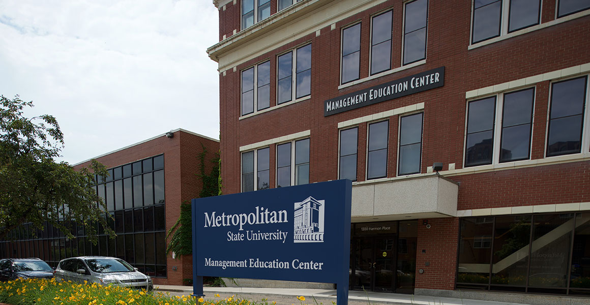 Photograph of Metro State University’s Management Education Center located in Minneapolis, Minnesota; red brick building with white trim, blue sign within an elevated bed of yellow lilies across the street but in front of the building stating, in white lettering, “Management Education Center” with Metro State’s logo shown in white.