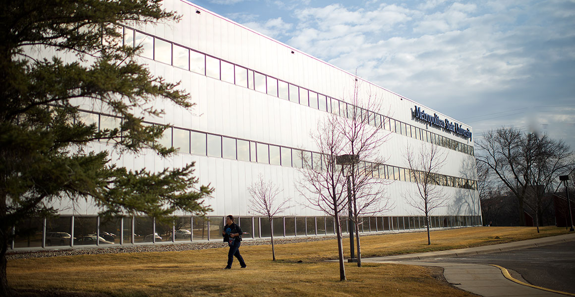 Photograph of the West side of Metro State University’s Midway Center in the very early Spring, with yellow grass and bare trees; a man walks on the sidewalk toward the building from the parking lot.