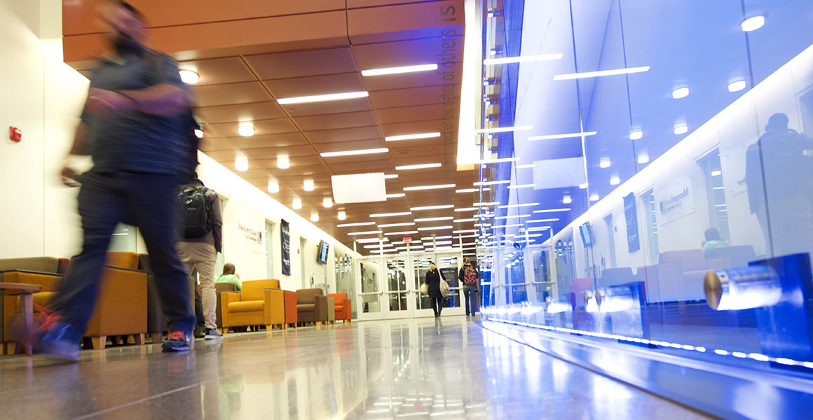 Photograph of students walking in a well-lit corridor at one of Metro State University’s locations; bright lights, reflects, and the blur of a student walking past.