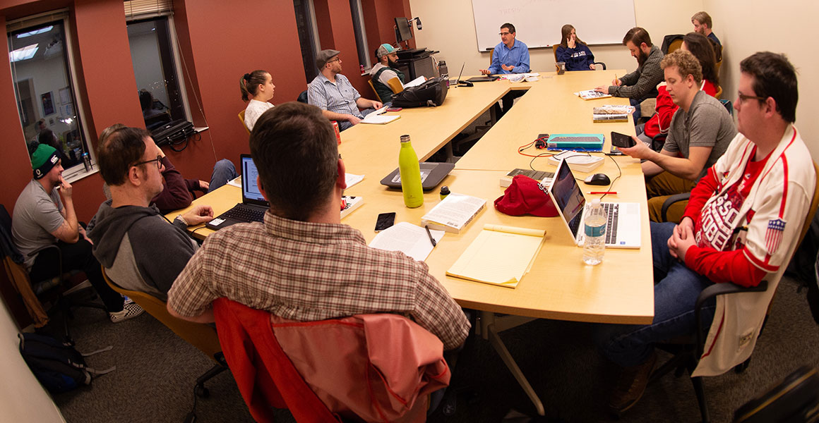 Photograph of students gathered around large tables in a smaller meeting room using laptops or taking notes as they enjoy a discussion with the professor in a more non-traditional environment at one of Metro State University’s teaching locations.