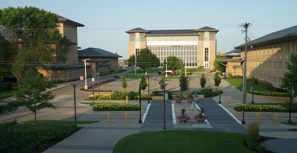 Photograph of Metropolitan State University’s Saint Paul, Minnesota campus, shot elevated from the East on an early Summer day overlooking Maria Avenue and the center courtyard parking area with flowers blooming, green grass and trees.