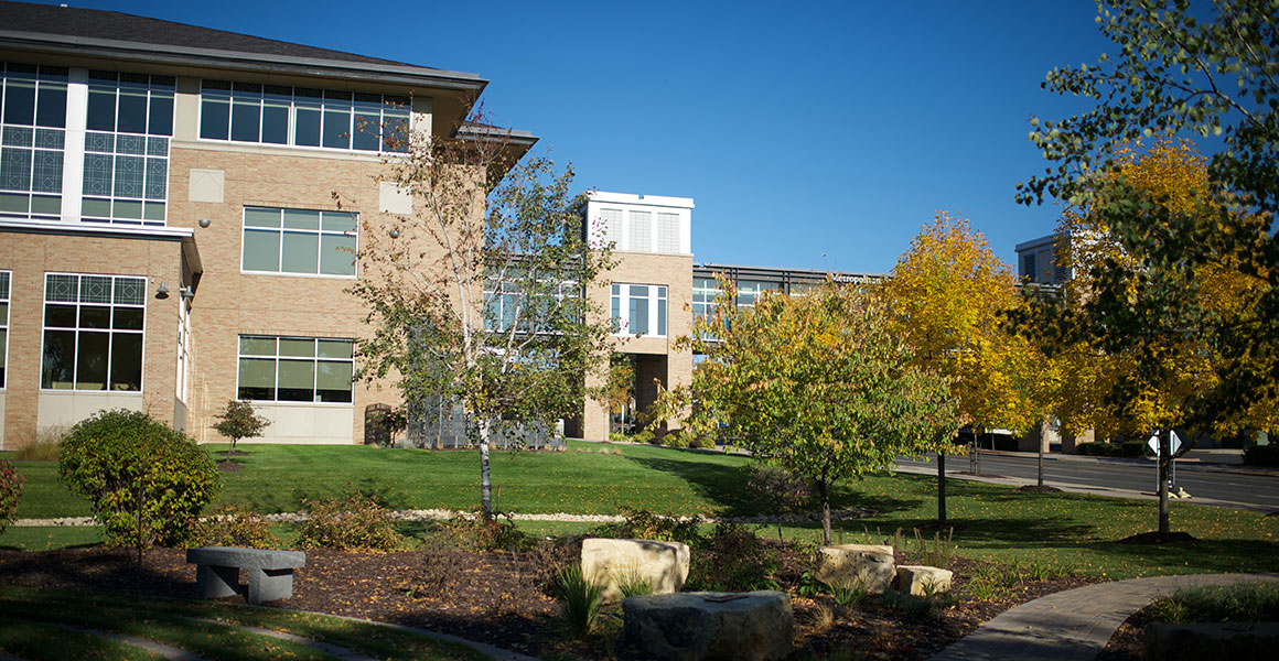 Photograph of Metro State University’s library and learning center taken from the labyrinth, West to East, on a late summer or early fall bright day with some leaves on the ground and trees turning from green to yellow.