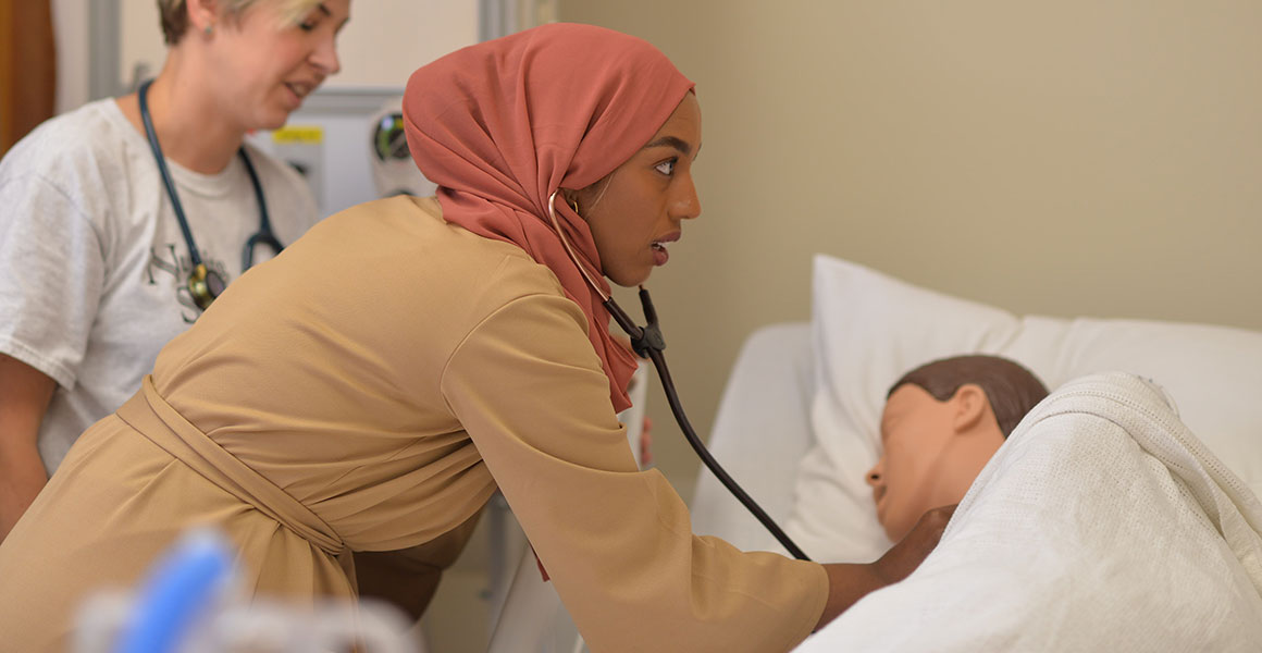 Photograph of a female nursing student wearing a coral colored hijab checking the pulse of a medical representation of a human subject lying in a hospital bed with the nursing instructor looking on.
