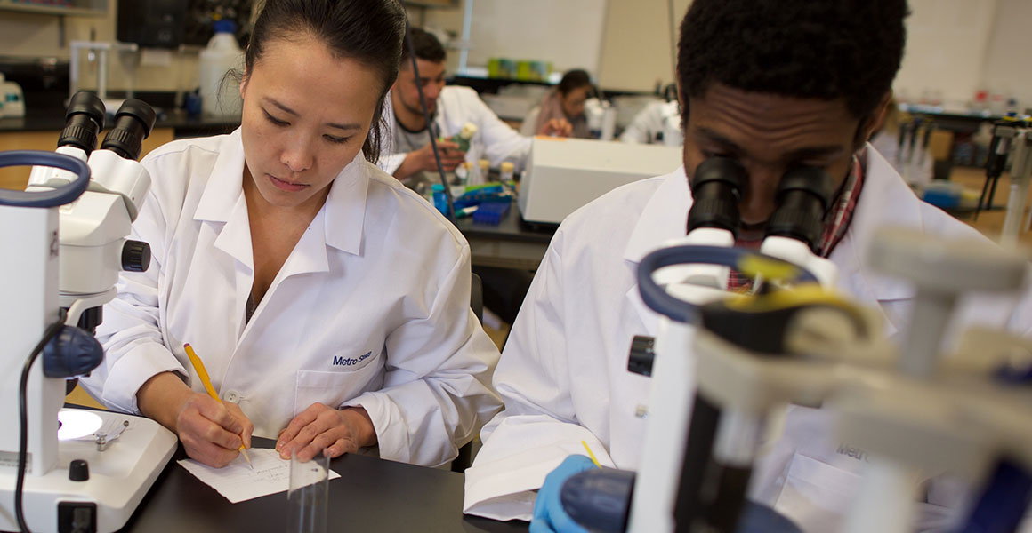 Photograph in a science lab in Metropolitan State’s Jason R. Carter Science Education Center on the Saint Paul, Minnesota Campus; an Asian American female student taking notes on the left working with an African American male student who is analyzing a slide in a microscope.