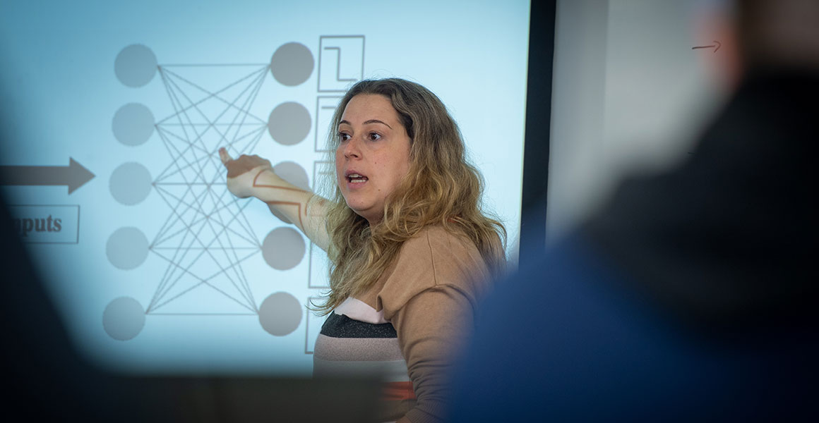 A female instructor stands in front of a projection; she points at black and white a diagram with two columns of circles and lines connecting the columns and various angles.