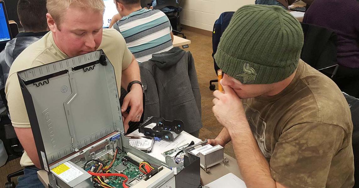 Two white men sit at a table with an open computer open in front of them; the electronic components of the computer are exposed.