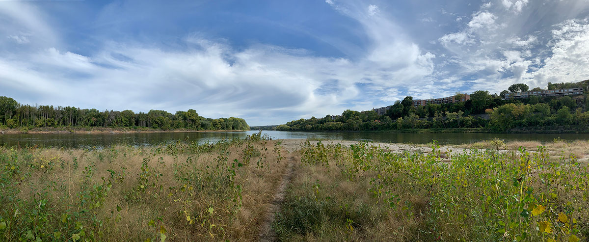 The confluence of the Minnesota and Mississippi Rivers; flat, wooded land is to the left, a grassy area is in front, and a wooded bluff with buildings overlooking the rivers is at the right.
