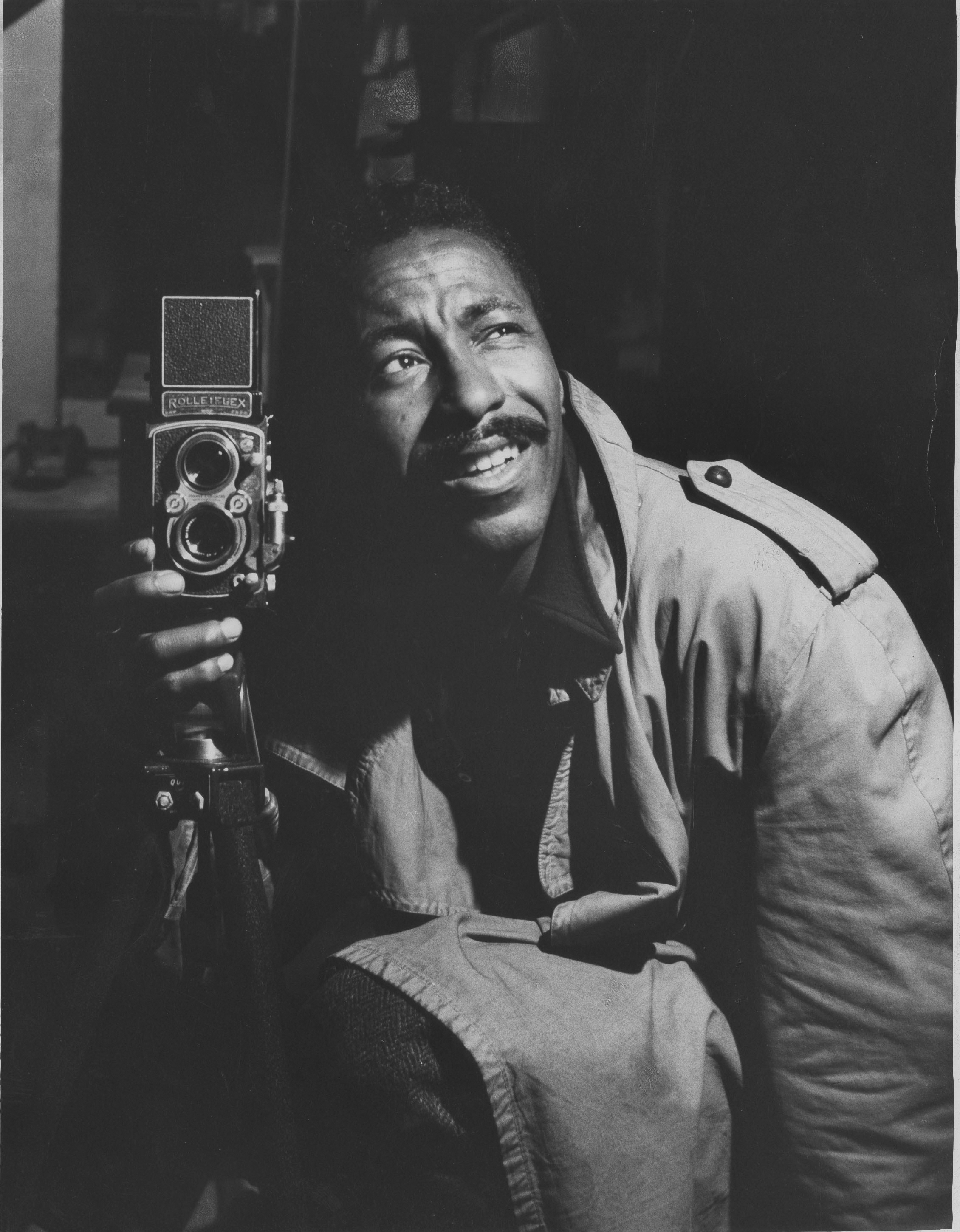 Picture of a young Gordon Parks in a trenchcoat, holding an old film camera