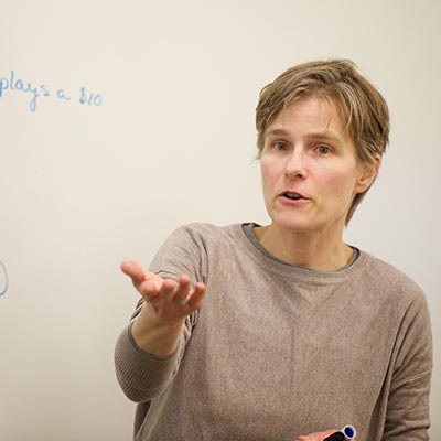 A woman lectures to a class in front of a dry-erase board.