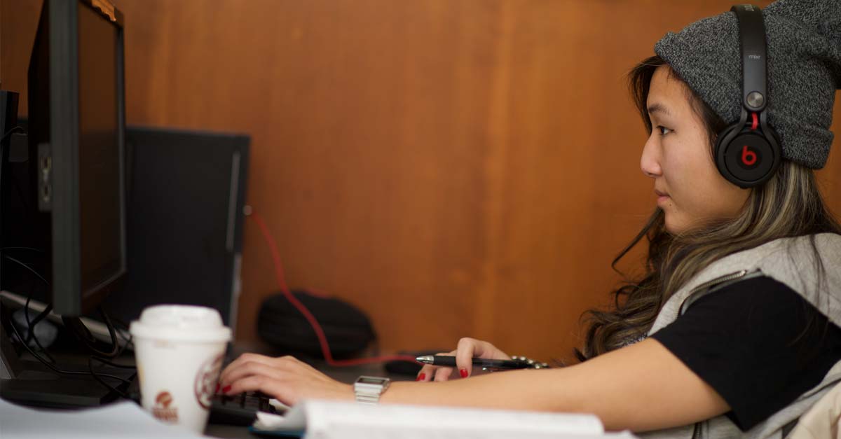 A young Asian woman wearing headphones and working at a laptop sits in a library.