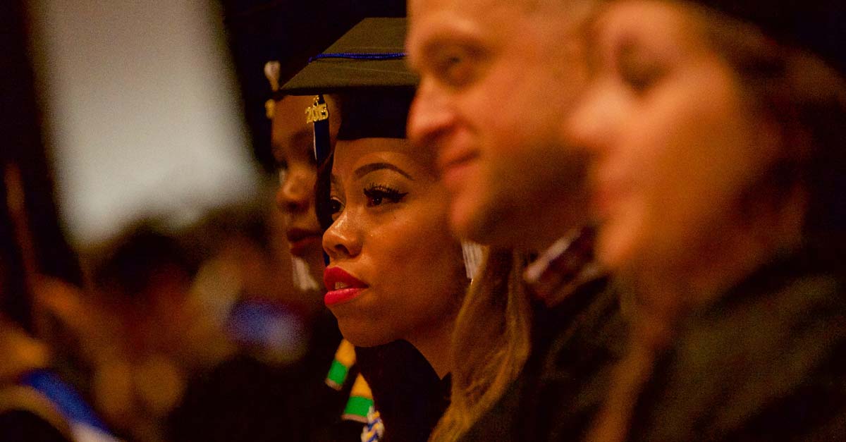 A group of graduates dressed in caps and gowns listen to a speaker outside of the image frame.