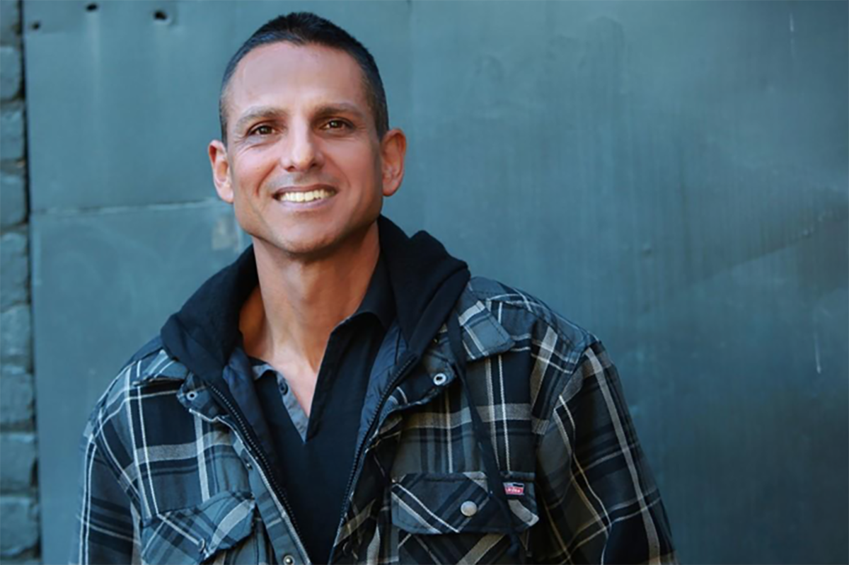 A man in a plaid jacket smiles as he stands in front of a blue-gray wall