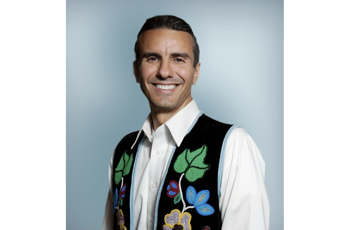 a man stands for a portrait in a white shirt with an intricately-beaded vest before a blue-gray backdrop