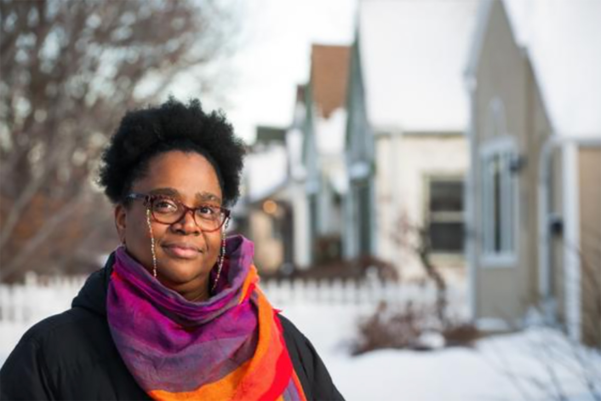 A woman wearing a colorful scarf stands in front of a row of houses