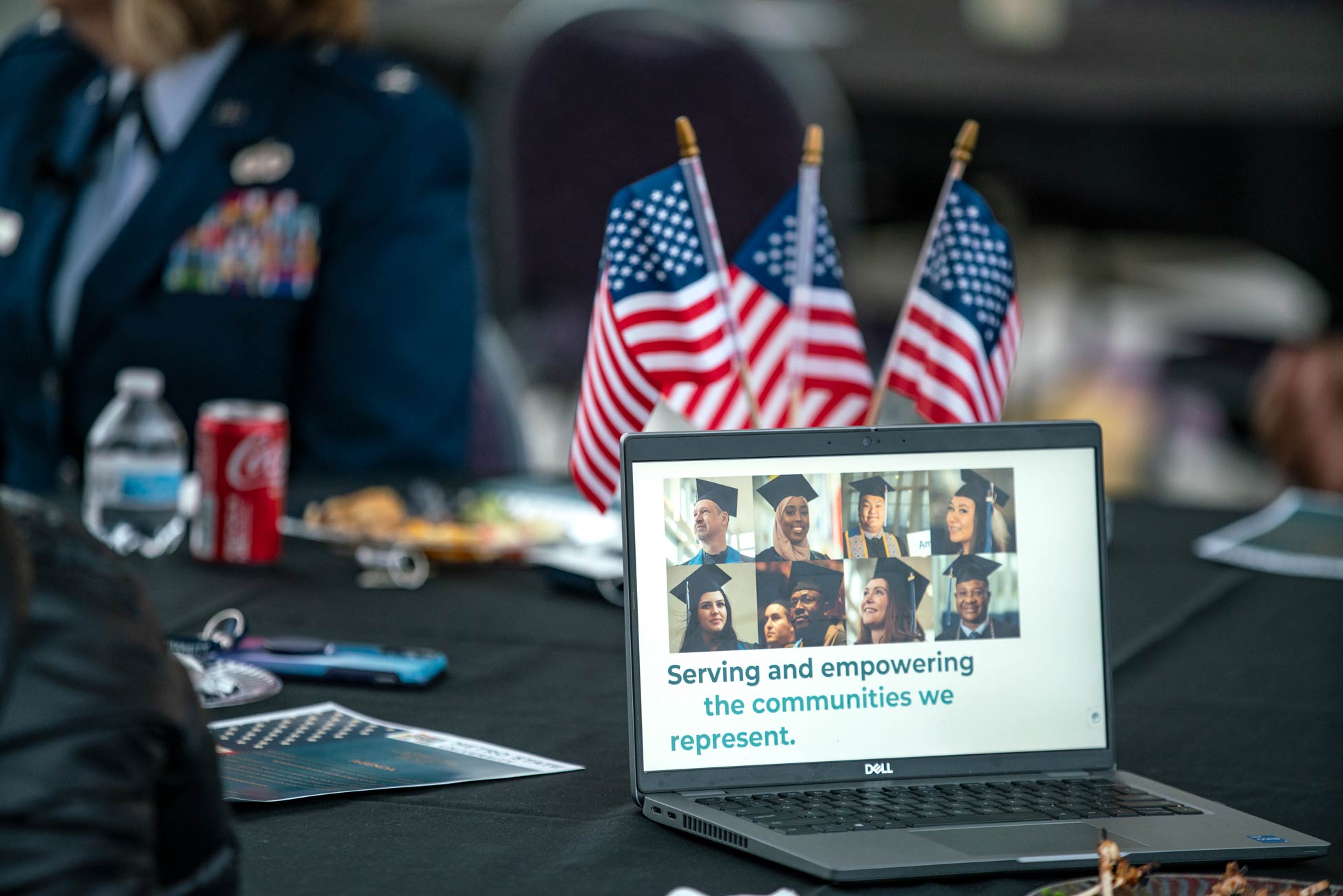 A laptop screen with faces of graduates sits on a table with flags and the message "serving and empowering the communities we represent" 