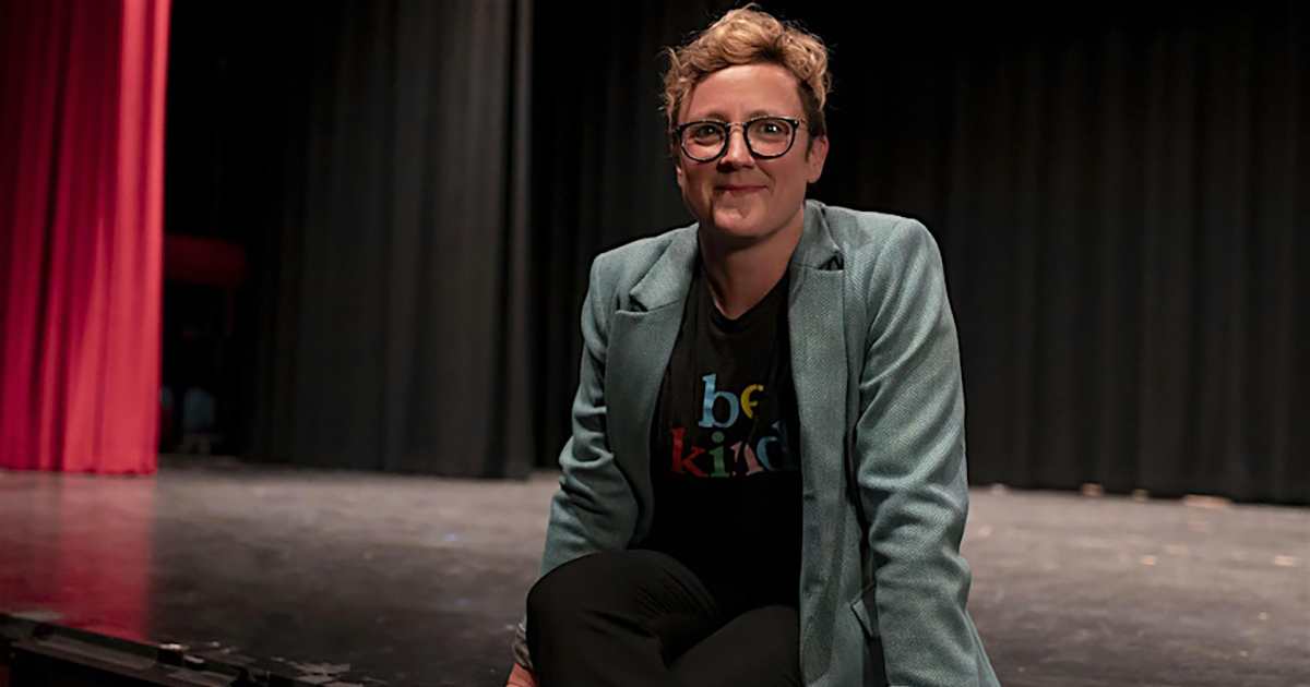 A person wearing glasses poses on the edge of a stage with a red curtain behind them