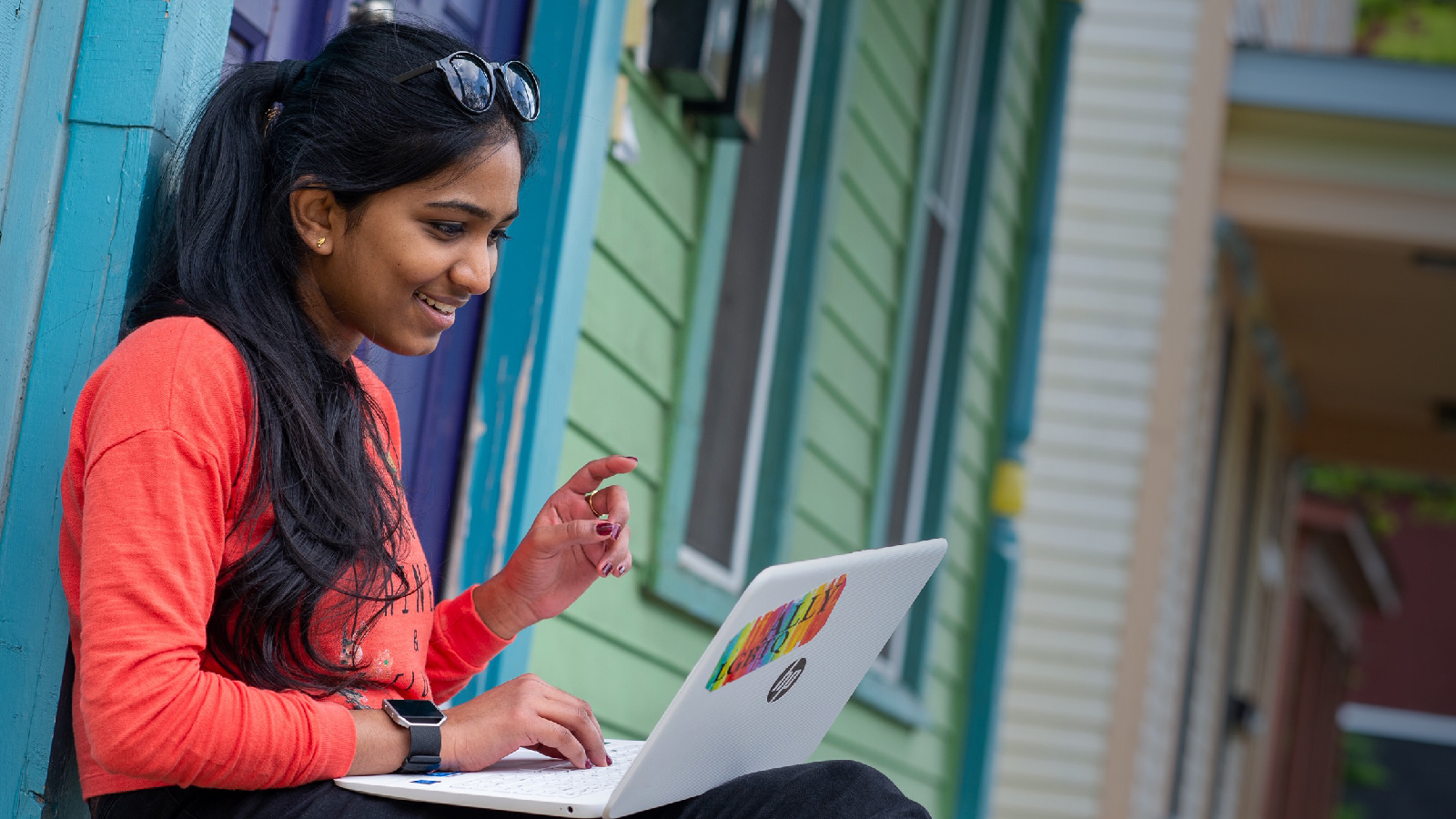 A woman works on a laptop while sitting in front of a colorful house