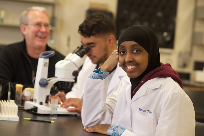 Two students in white lab coats in lab with microscope and faculty
