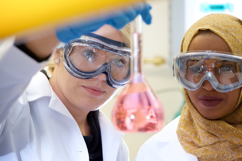 two female scientists in lab coats and safety goggles with an experiment