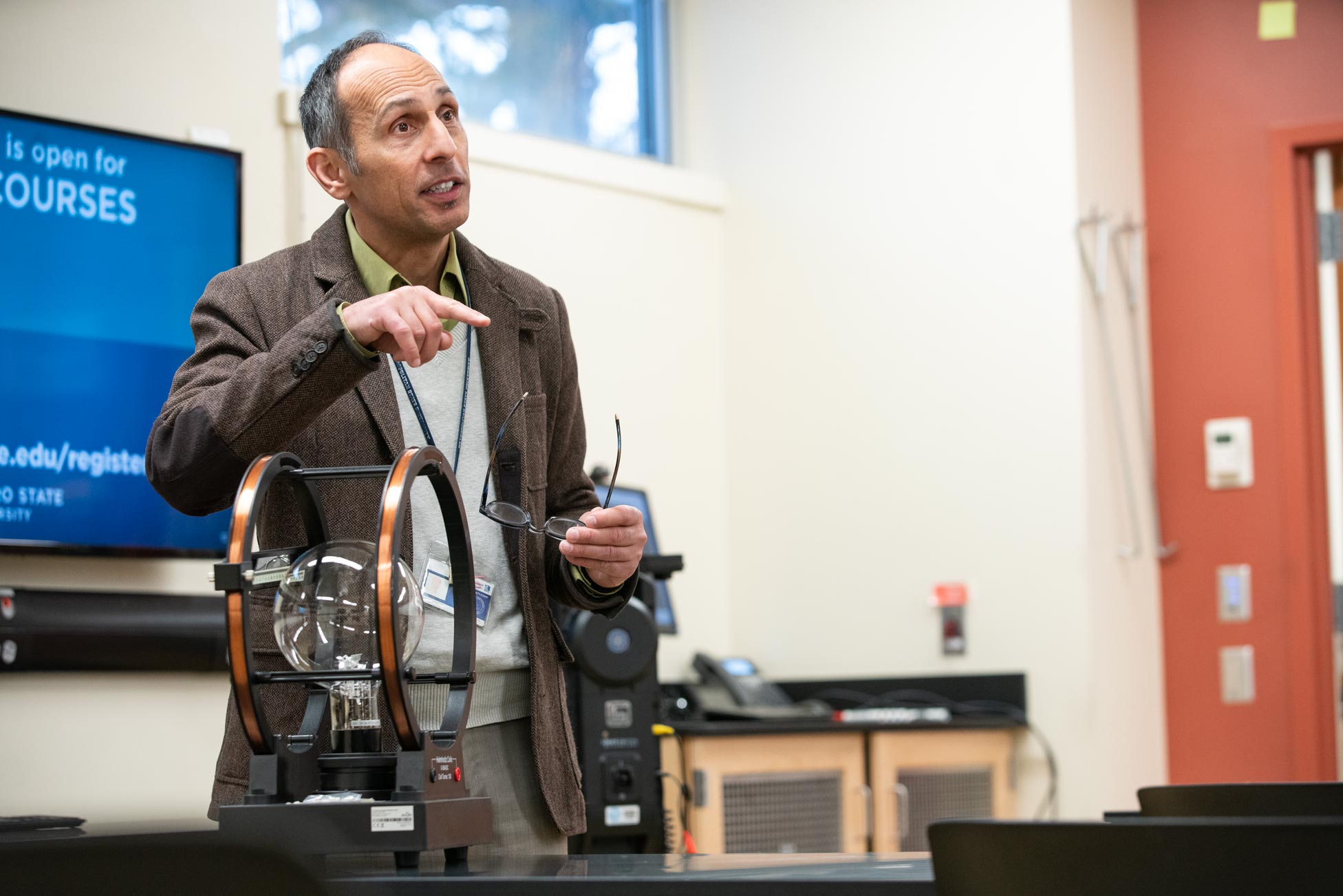 A man in a brown jacket lectures in front of a screen and near a piece of lab equipment