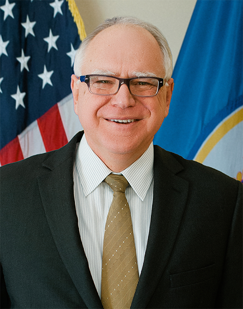 Governor Tim Walz in a dark suit stands before U.S. and State of Minnesota flags
