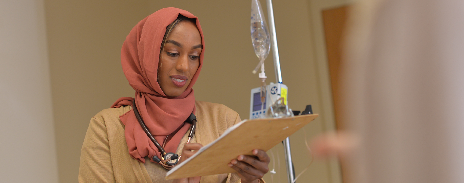 Female nursing student wearing a a hijab, reviewing a chart while standing next to an IV unit