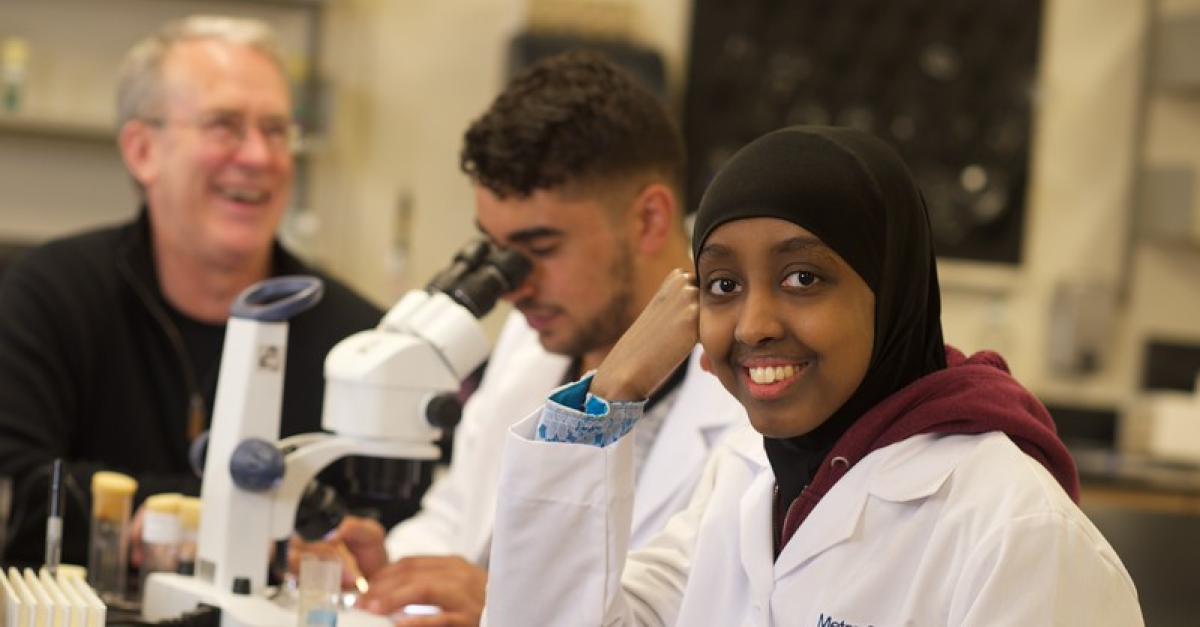 Two students in white lab coats in lab with microscope and faculty