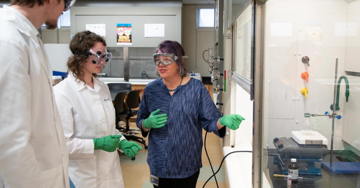 An instructor in safety gear explains a chemistry lab station to two students in safety gear