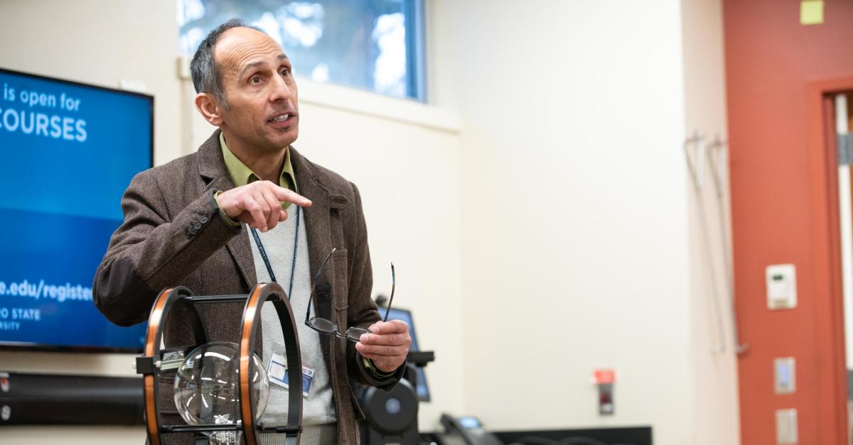 A man in a brown jacket lectures in front of a screen and near a piece of lab equipment