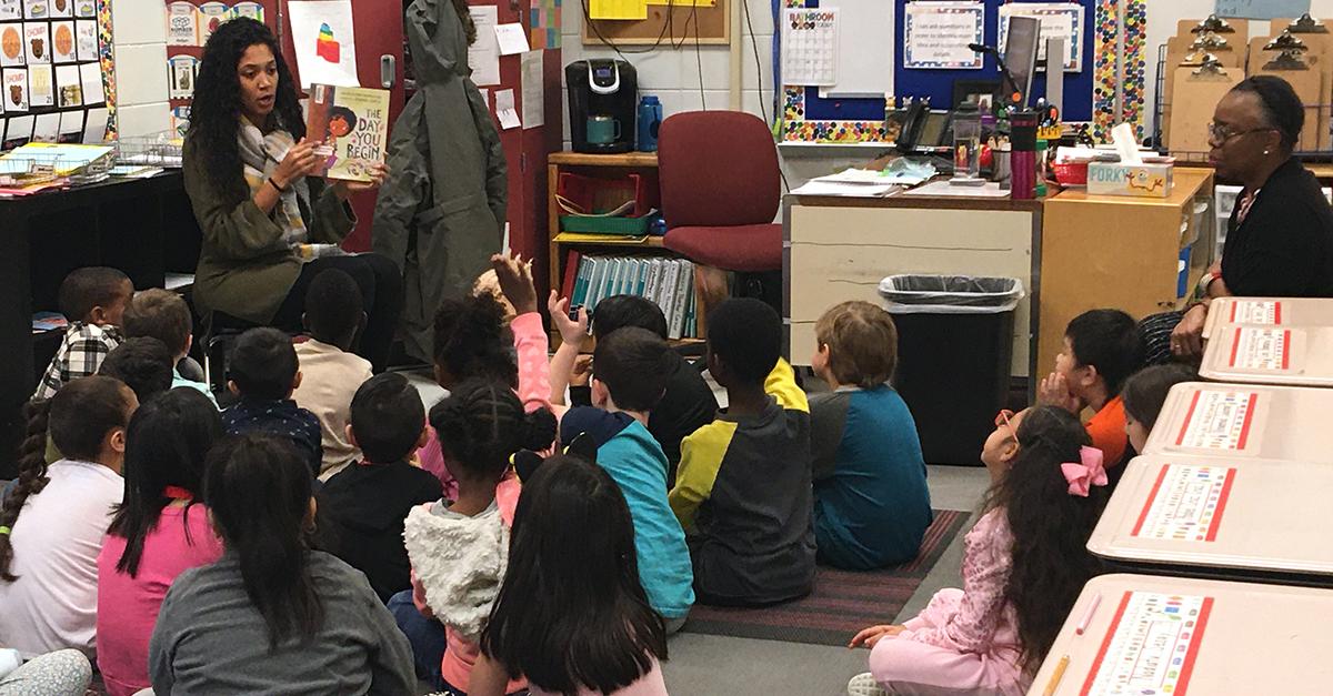 Teacher-in-training reading a book to a group of children in their classroom with professor looking on
