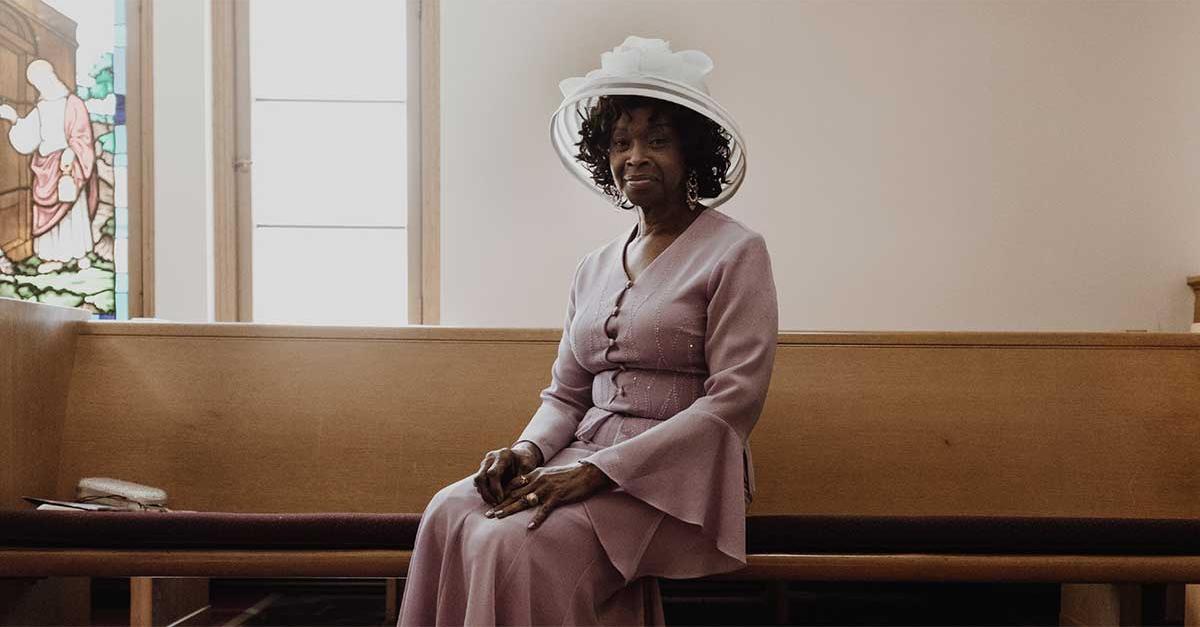 An African-American woman wearing a pink dress and white hat sits in a church pew.