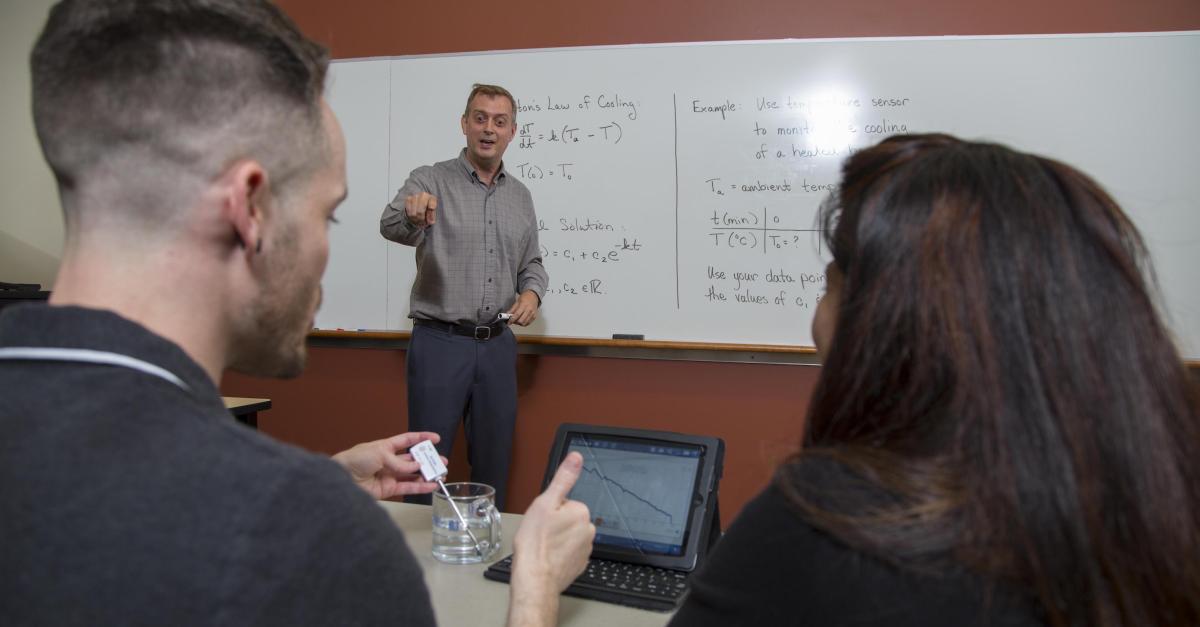 Math faculty at whiteboard speaking to two students