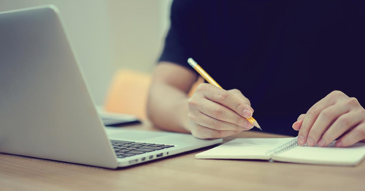 person writing in a notebook and an open laptop on desk