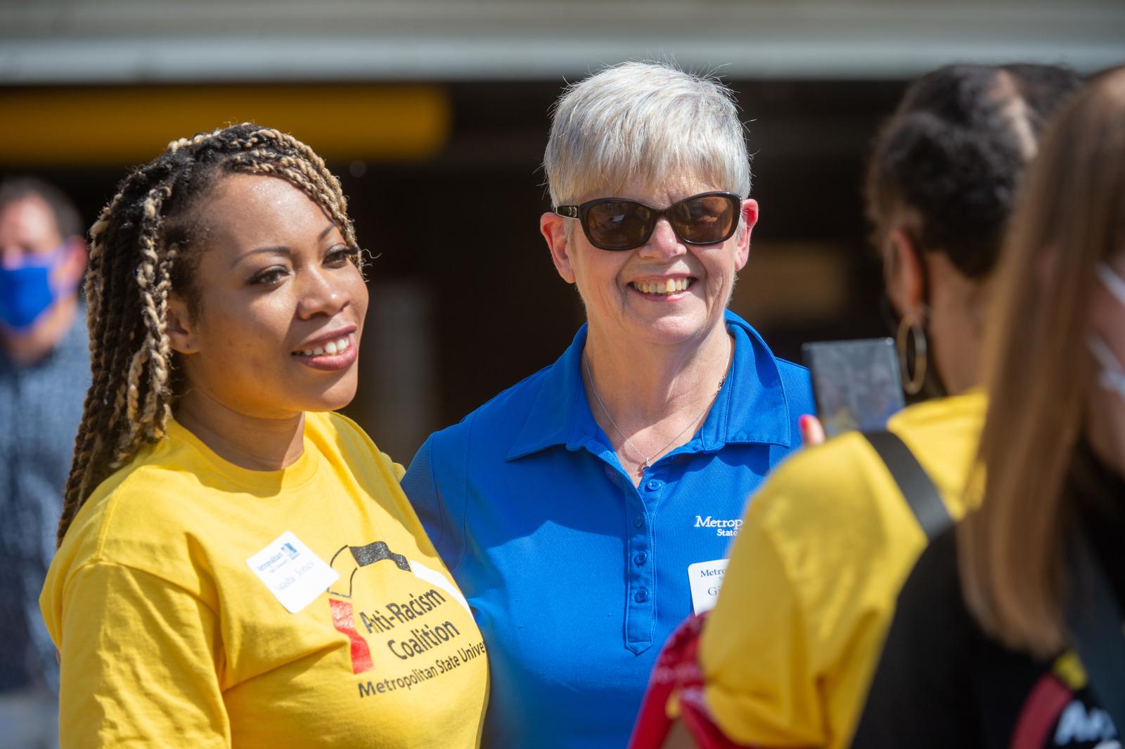 two women stand shoulder to shoulder smiling towards others