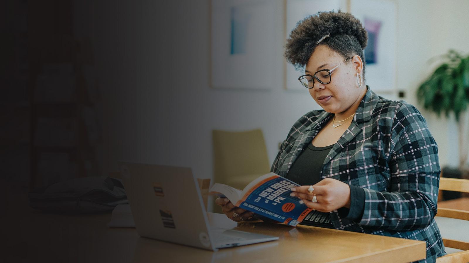A student reading a text book while studying at a laptop