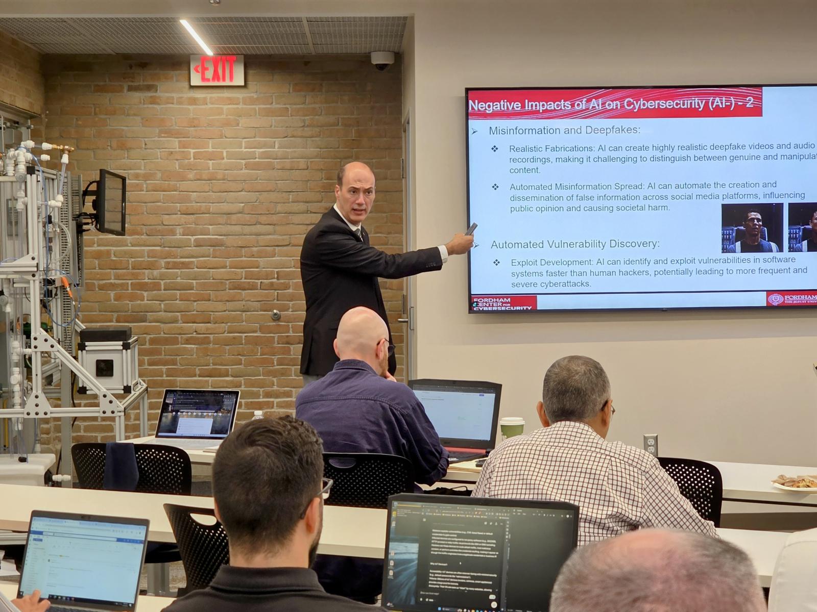 A person at a wall monitor teaching a classroom of people at laptops