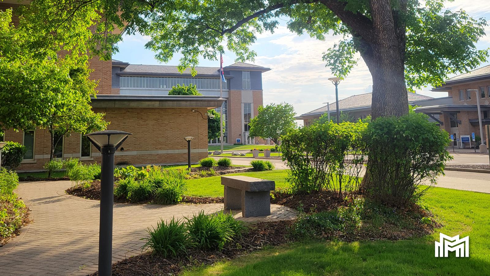 Metro State campus seen from the walking path near Founders Hall, with the tree and stone bench in the foreground