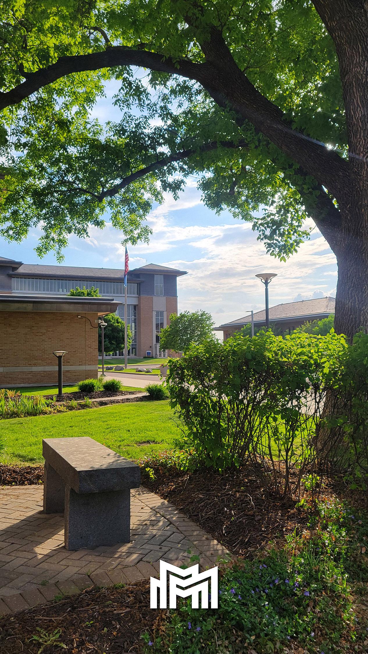 Metro State campus framed by a tree bower with a stone bench in the foreground, and the Metro State icon in white