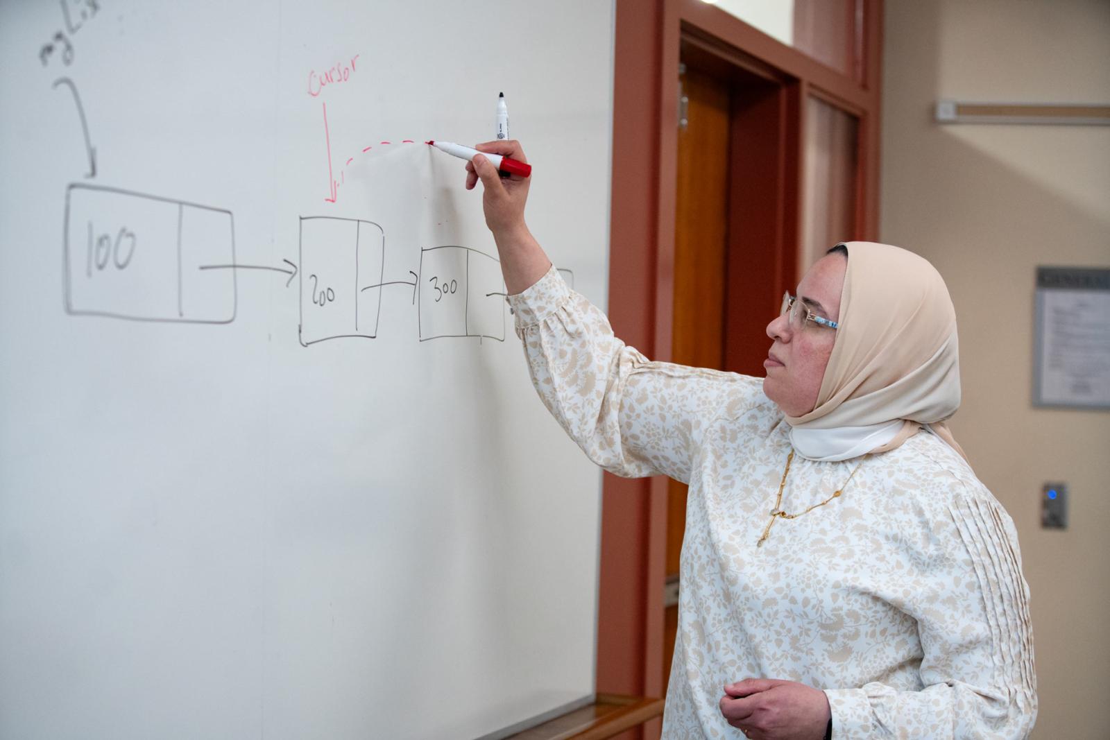 an instructor with a light headscarf writes at a whiteboard