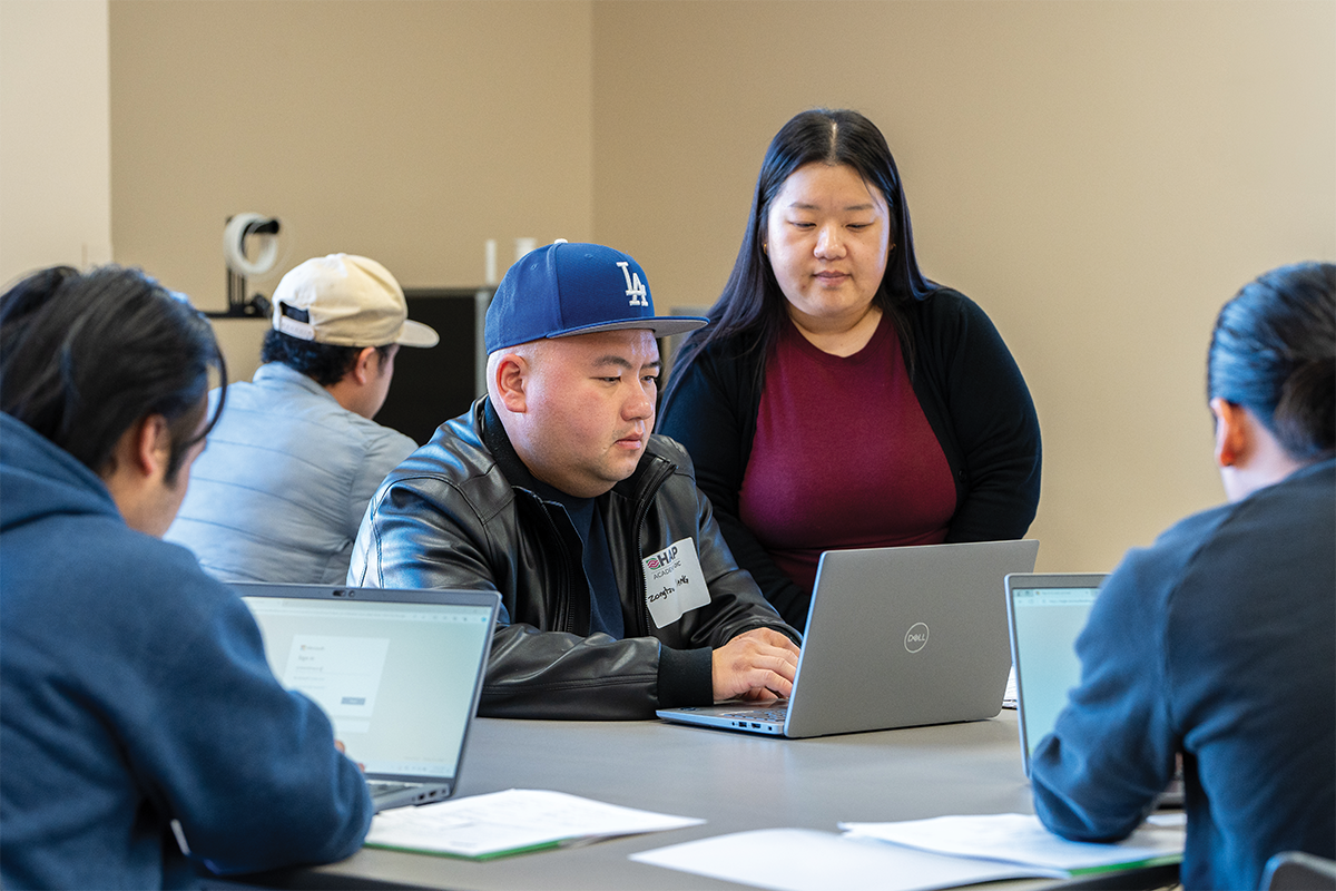 A group of people working in a classroom at laptops, with an instructor helping one of them