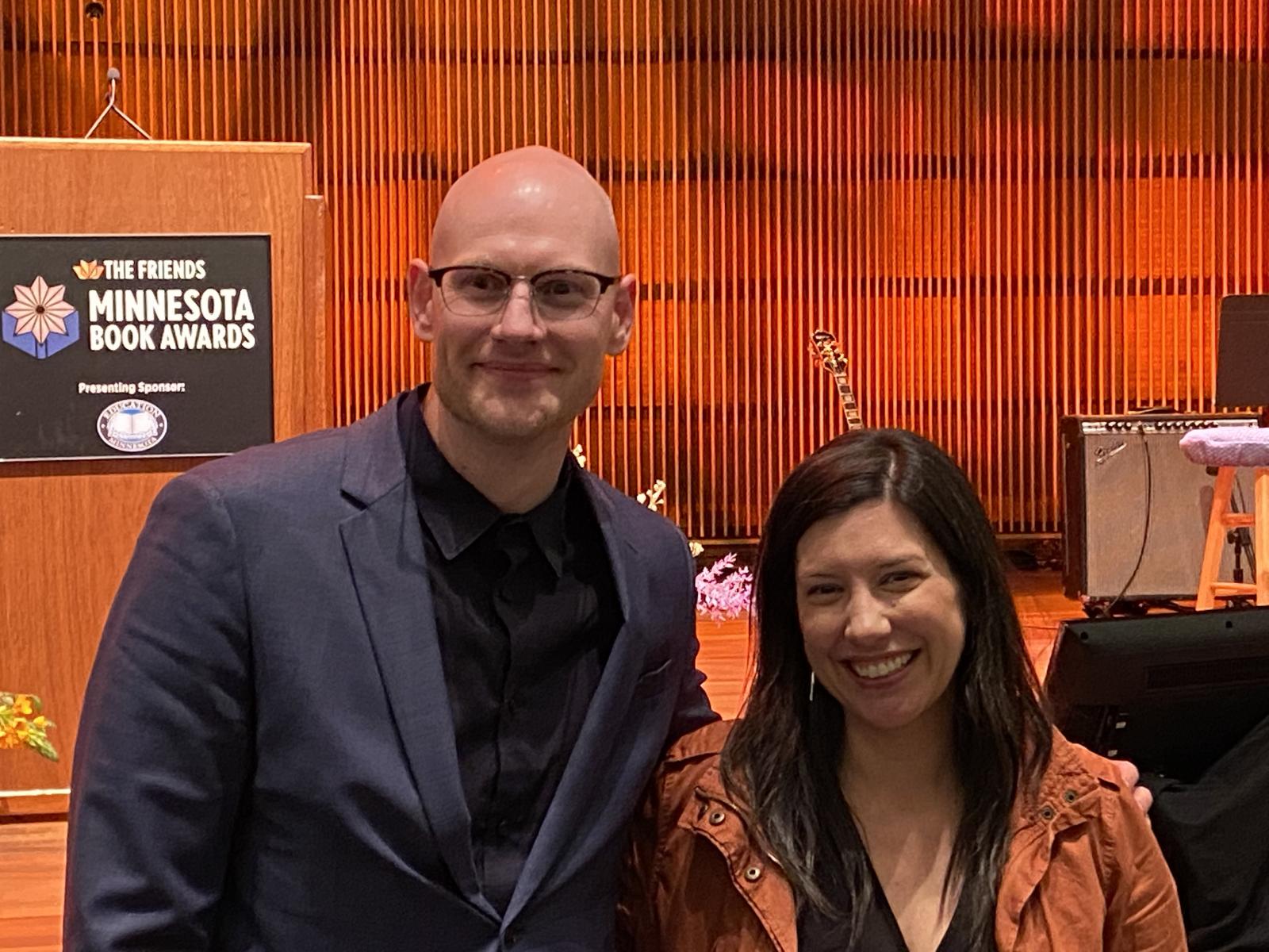 Two people pose for a photo in front of a lecturn, a guitar, and some sound equipment before an orange stage wall