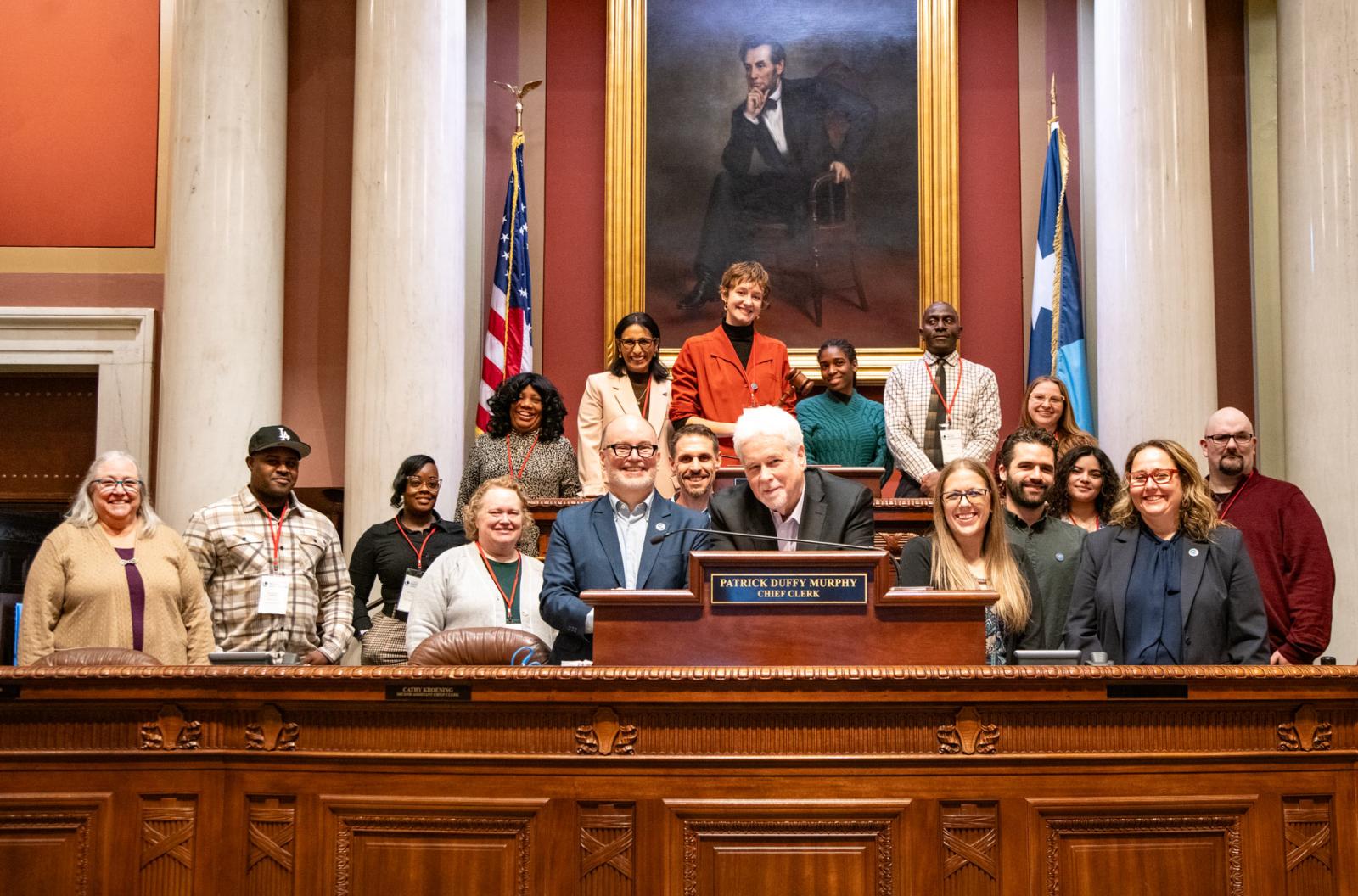 A group pf people pose for a group portrait at the Minnesota State House of Representatives Speaker's Dais