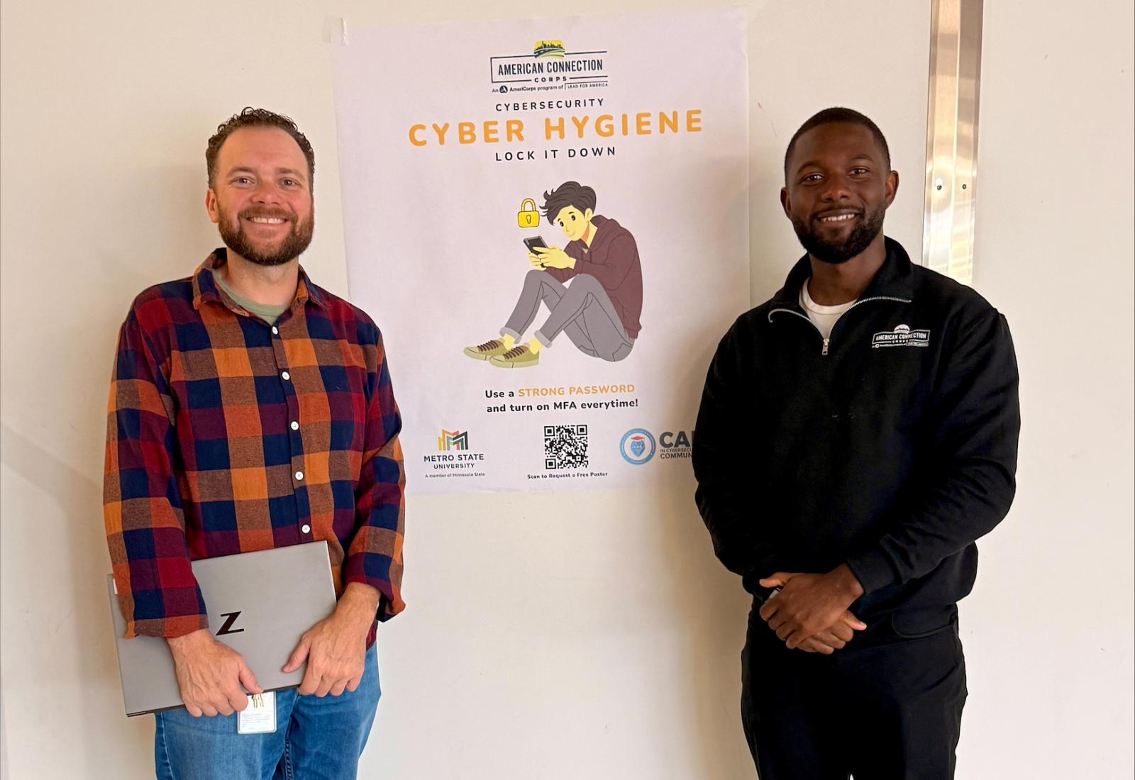 Two men stand posing for a photo, one holding a laptop, in front of a poster for a cyber hygiene clinic
