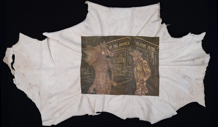 A screen print of a person with a horse head wearing a dress and a man with a hat holding a bottle standing outside the corner of a building with a words "De Mis Amores, El Amor De Mis" printed in sepia tones on a roughly torn out scrap of dirty white fabric