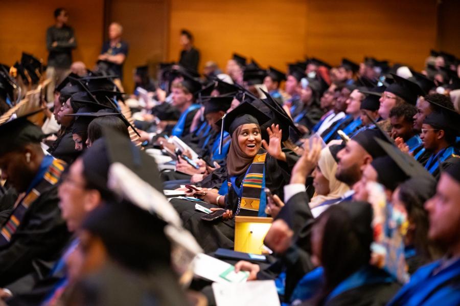 A woman in cap and gown waves to someone while sitting in rows of fellow students at commencement