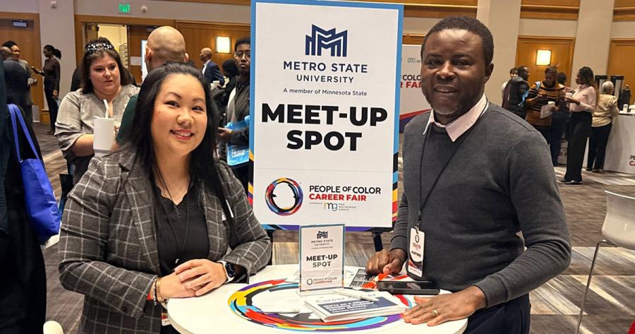 a woman and man standing next to a cafe table with Metro State Meet-up spot: People of Color Career Fair on a sign on top. 