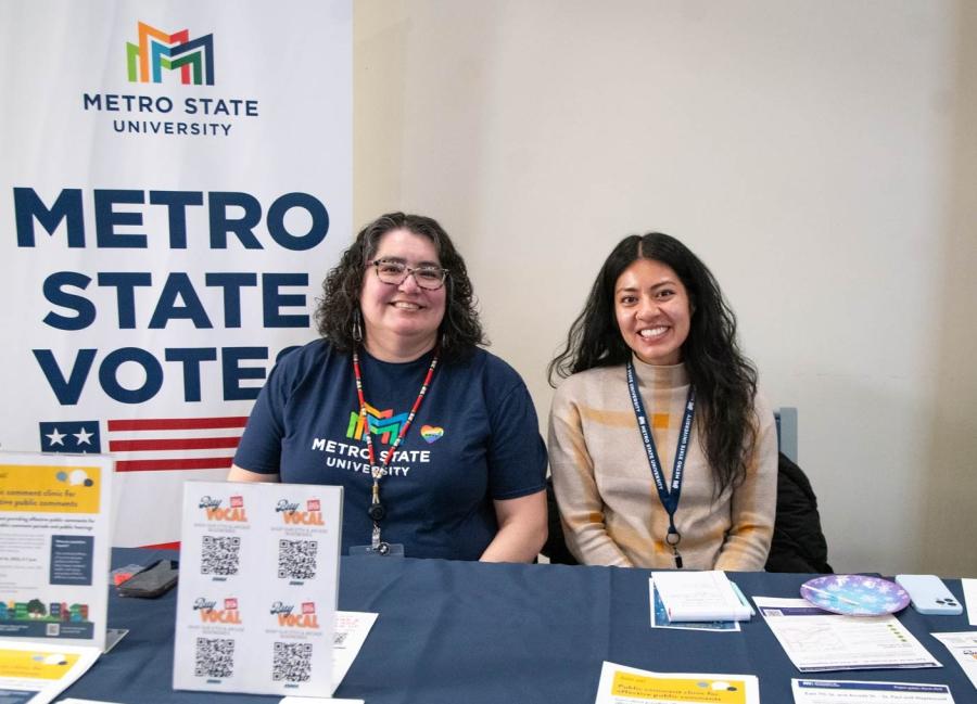 Two people pose behind a table in front of a Metro State Votes banner